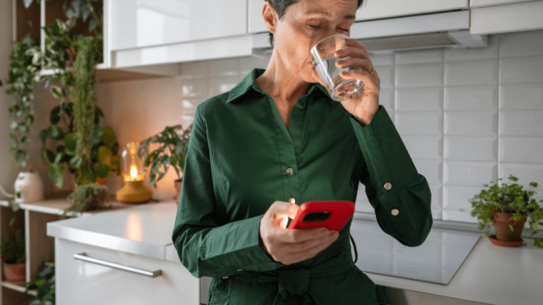 Kvinna i kök som scrollar på en mobiltelefon samtidigt som hon dricker vatten